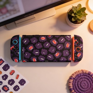 Puede incluir: Una consola Nintendo Switch negra con un patrón de fantasmas morados de dibujos animados. Los mandos son negros y naranjas con diseños de fantasmas a juego. Se ve una hoja de pegatinas con el mismo diseño.