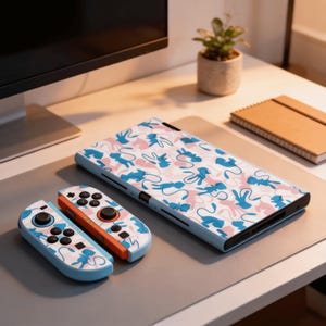 Peut inclure: Une console Nintendo Switch bleu clair et des manettes avec un motif répété de personnages de dessins animés bleus et roses. Les manettes ont des boutons noirs et des accents orange. La console et les manettes sont sur une surface grise.