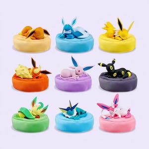 May include: Nine colorful plush figures of Pokémon characters resting on small, round cushions. The figures include Eevee, Glaceon, Jolteon, Flareon, Espeon, Umbreon, Leafeon, Vaporeon, and Sylveon, each with unique colors and designs.