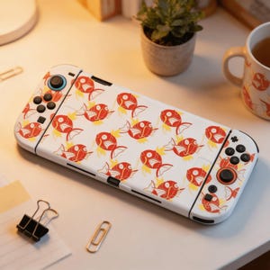 Peut inclure: Une console Nintendo Switch blanche ornée d'un motif répété de poissons rouges et jaunes. La console est posée sur une surface blanche avec une petite plante en pot et une tasse en arrière-plan. Un trombone et une pince à reliure sont également visibles.