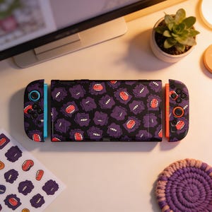 Peut inclure: Une console Nintendo Switch noire avec un motif de fantômes violets. Les manettes sont noires et oranges avec des motifs de fantômes assortis. Des autocollants avec le même motif sont également visibles.