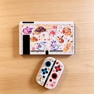 May include: White Nintendo Switch console and Joy-Cons featuring a colorful illustration of various cartoon characters. The console has a decorative border and the Joy-Cons have blue and red buttons.