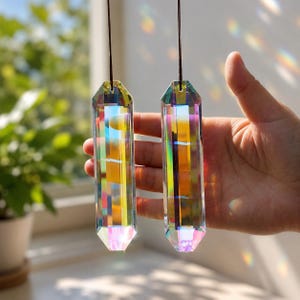 Pode incluir: Dois prismas de vidro hexagonais alongados com um acabamento transparente e iridescente que reflete um espectro de cores do arco-íris. Cada prisma apresenta bordas facetadas e está suspenso por um cordão fino castanho escuro.