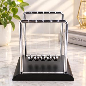 Peut inclure: Un pendule de Newton composé de cinq sphères métalliques argentées polies, suspendues par de fins fils à un cadre chromé. L'ensemble est fixé sur une base rectangulaire noire solide, formant un dispositif physique équilibré et symétrique.
