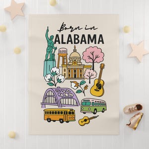 May include: A cream-colored blanket featuring a colorful illustration of Alabama landmarks, including a statue, capitol building, and guitar. The text "Born in Alabama" is displayed in a handwritten font.