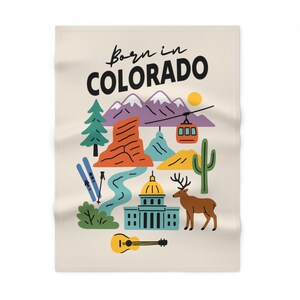 Puede incluir: Manta de color crema con una colorida ilustración de los lugares emblemáticos de Colorado, incluyendo montañas, un teleférico y un ciervo. El texto "Born in COLORADO" está en la parte superior en blanco y negro.