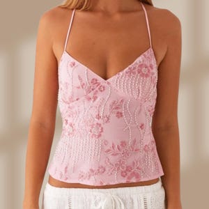 May include: A light pink halter top with a V-neckline. The top is adorned with floral embroidery and beaded accents. The garment has a delicate, feminine design, suitable for warm weather or special occasions.