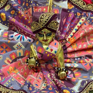 May include: A gold deity statue wearing a turban and ornate clothing. The statue is dressed in vibrant purple, pink, and gold fabrics, featuring detailed patterns and embellishments. The design is highly decorative, with a focus on rich colours and textures, typical of religious iconography.