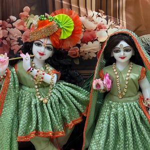 May include: Two statues of deities, adorned in green and orange attire. One holds a flute, the other a flower. Both wear elaborate jewellery and headdresses. The backdrop includes floral and patterned designs.