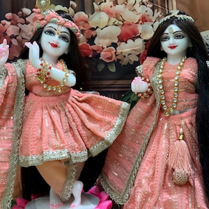 Peut inclure: Deux statues de divinités, vêtues de vêtements traditionnels pêche avec des bordures dorées. Les statues ont des cheveux noirs, des coiffes élaborées et des bijoux. L'une des statues est posée avec les mains en geste, l'autre se tient à côté. Une plume de paon orne la tête d'une statue.