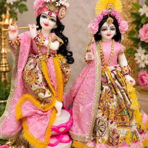 May include: Two ornate figurines of deities dressed in matching pink and yellow outfits with intricate patterns. The left figure holds a gold flute, while both wear elaborate floral headpieces, beaded necklaces, and gold-trimmed fabric garments.