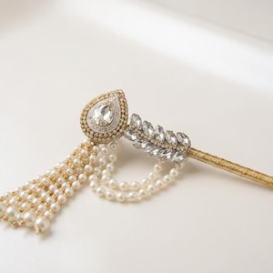 Puede incluir: Un accesorio decorativo para el cabello con un tallo dorado, un cristal central en forma de lágrima rodeado de pequeñas cuentas blancas, un diseño de hojas con cristales transparentes y múltiples hilos colgantes de pequeñas perlas blancas.