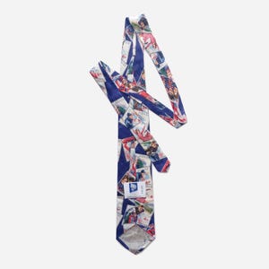May include: A navy blue necktie featuring a repeating pattern of vintage postage stamps depicting winter sports scenes. The stamps include illustrations of skiers and skaters with the text 4kr NORGE. A white label is attached to the front.
