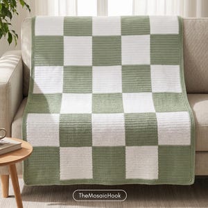 May include: A rectangular crochet blanket featuring a repeating checkerboard pattern in alternating sage green and white squares. The textile has a consistent stitch texture throughout and is finished with a simple border in the same sage green yarn.