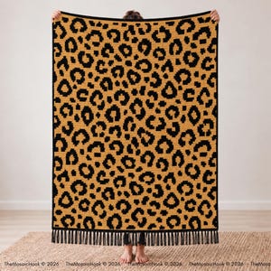 Puede incluir: Una manta rectangular de ganchillo con un patrón repetitivo de estampado de leopardo negro sobre un fondo amarillo dorado. La manta está terminada con un borde negro sólido y flecos de hilo negro a lo largo del borde inferior.