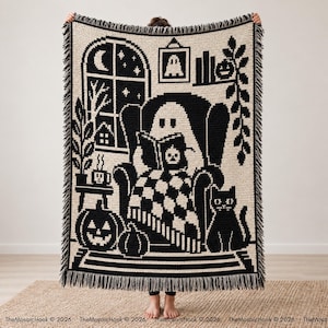 Pode incluir: Uma manta tecida em preto e creme com uma ilustração pixelizada de um fantasma lendo um livro numa poltrona, um gato preto, abóboras, uma janela com uma lua crescente e um quadro de fantasma, com acabamento em franjas.
