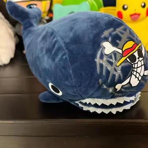May include: A plush, navy blue whale toy with a white-toothed mouth and a pirate-themed design on its side. The design features a skull wearing a yellow hat with a red band. Other colorful plush toys are in the background.