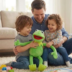 May include: A plush green Kermit the Frog puppet with a red mouth and yellow collar is held by two children and an adult. The puppet is the focal point, with the children and adult smiling. The scene is set indoors on a light-colored carpet.