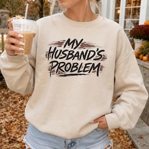 May include: A beige crewneck sweatshirt featuring the black text "MY HUSBAND'S PROBLEM" printed over a distressed brown brushstroke graphic on the chest. The garment has long sleeves, a ribbed neckline, and ribbed cuffs, providing a casual look.