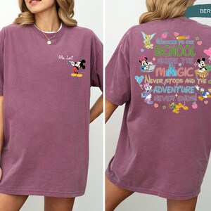 May include: Berry-colored t-shirt featuring Mickey Mouse on the front and a colorful graphic on the back. The back graphic includes the text "Welcome to our school where the magic never stops and the adventure never ends!" with various Disney characters.