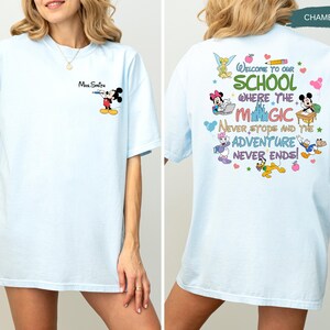 May include: Light blue t-shirt with a cartoon graphic of Mickey Mouse and the text "Mrs. Smith" on the front. The back of the shirt features a colorful design with the text "Welcome to our school where the magic never stops and the adventure never ends!"