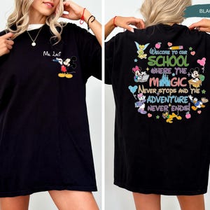 May include: Black t-shirt featuring Mickey Mouse on the front and a colorful graphic on the back that reads "Welcome to our school where the magic never stops and the adventure never ends." The shirt has a white collar.