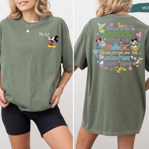 May include: Moss green t-shirt featuring Mickey Mouse with a water gun on the front. The back of the shirt has a colorful graphic with Disney characters and the text "Welcome to the School Where the Magic Never Stops and the Adventure Never Ends!"