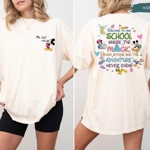 May include: Ivory-colored t-shirt featuring Mickey Mouse as a teacher with the text "Ma Lact" on the front. The back of the shirt displays the phrase "Welcome to our school where the magic never stops and the adventure never ends!" with Disney characters.