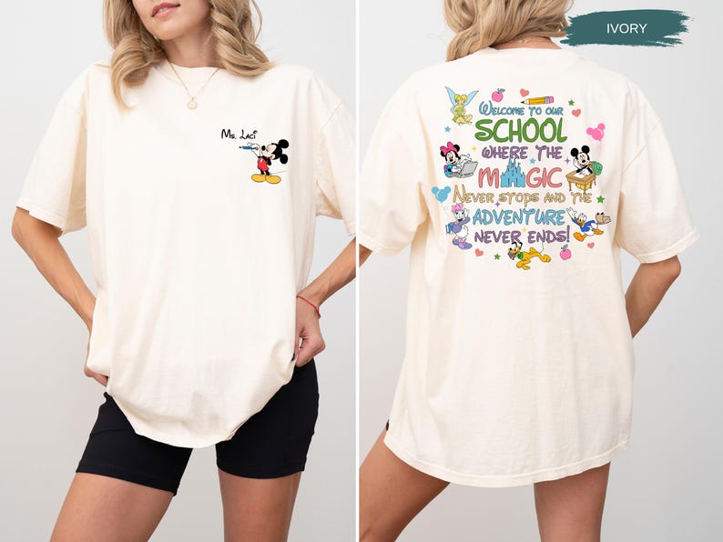 May include: Ivory-colored t-shirt featuring Mickey Mouse as a teacher with the text "Ma Lact" on the front. The back of the shirt displays the phrase "Welcome to our school where the magic never stops and the adventure never ends!" with Disney characters.