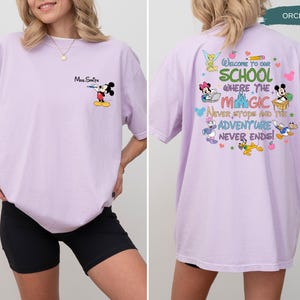 May include: Orchid-colored t-shirt featuring Mickey Mouse with a water gun on the front. The back of the shirt has a colorful graphic with Disney characters and the text "Welcome to our school where the magic never stops and the adventure never ends!"
