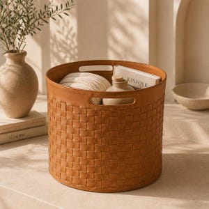 Op de afbeelding: Een ronde, tan opbergmand met een geweven buitenpatroon en twee geïntegreerde handgrepen. De mand bevat een wit textiel met textuur, een beige keramische fles met een houten stop en een boek met het woord KINFOLK op de kaft.