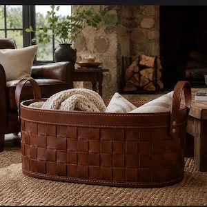 May include: An oval-shaped storage basket constructed from woven brown leather strips. The basket features two upright leather handles secured with metal rivets on each side. Inside the basket, a cream-colored knit blanket is folded neatly.
