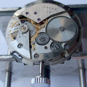 May include: Close-up of a watch movement, showcasing intricate gears, screws, and other components. The metal parts are silver and gold, with the text "2409A SU" visible on a plate. The watch is held in a metal clamp.