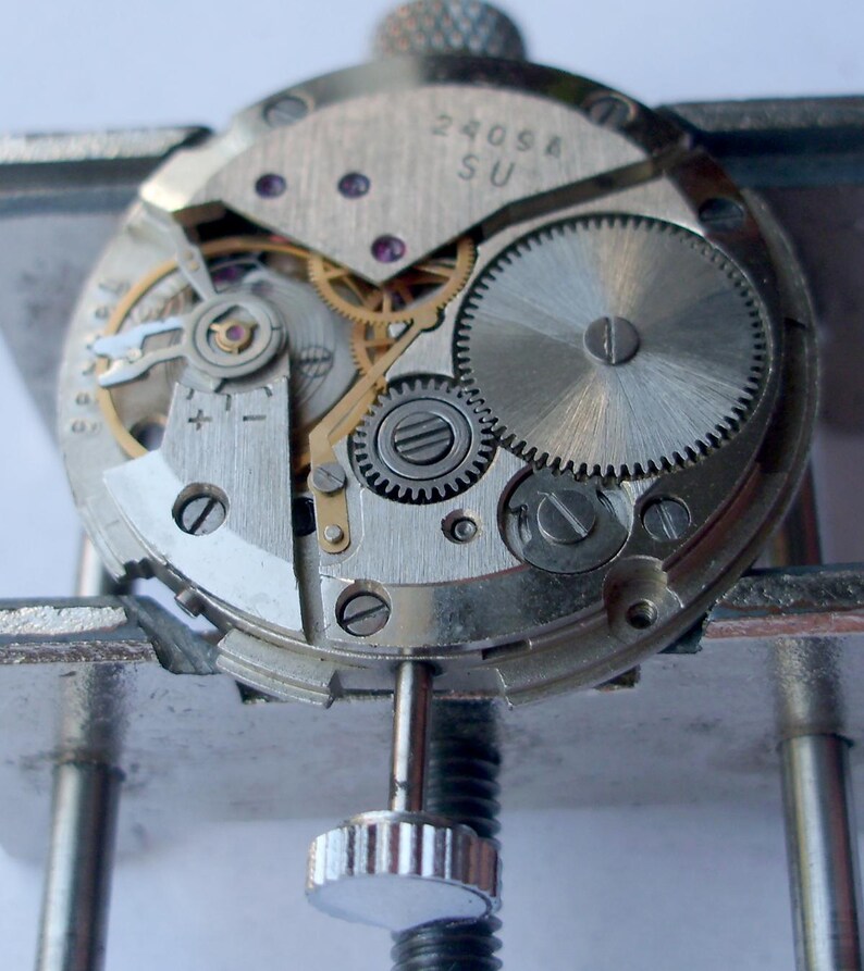 May include: Close-up of a watch movement, showcasing intricate gears, screws, and other components. The metal parts are silver and gold, with the text "2409A SU" visible on a plate. The watch is held in a metal clamp.