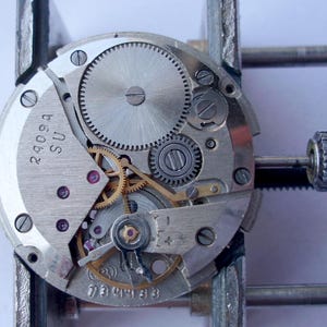 Może przedstawiać: Zbliżenie mechanizmu zegarka z misternymi kołami zębatymi, sprężynami i śrubami. Metalowe elementy są srebrne i złote, z widocznymi napisami "2909A SU" i "739433". Mechanizm zegarka jest trzymany w metalowym zacisku.