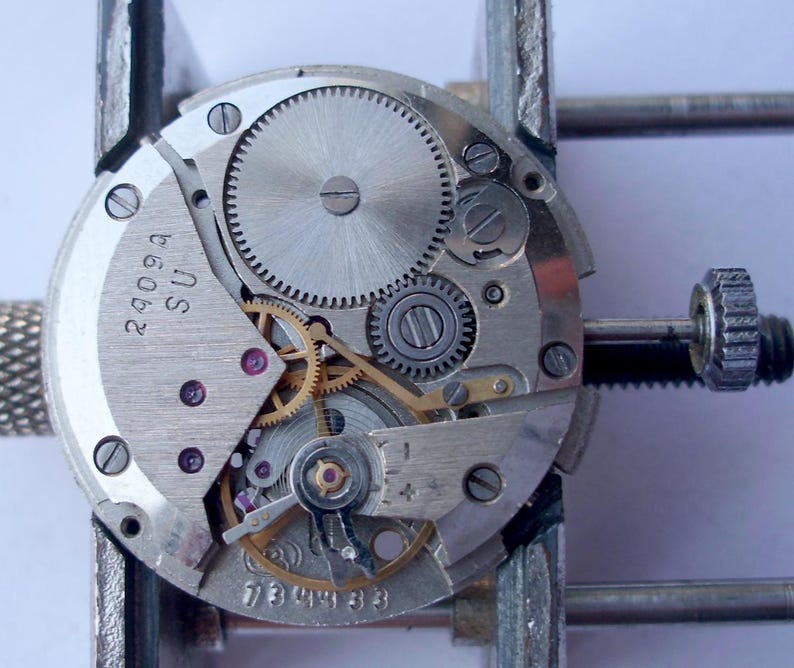 May include: Close-up of a watch movement with intricate gears, springs, and screws. The metal components are silver and gold, with the text "2909A SU" and "739433" visible. The watch mechanism is held in a metal clamp.