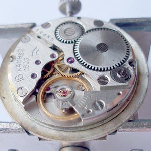 May include: Close-up of a disassembled watch movement. The silver-colored mechanism features numerous gears, screws, and jewels. The watch's internal components are visible, including the balance wheel and escapement. The watch movement is held in a silver metal frame.