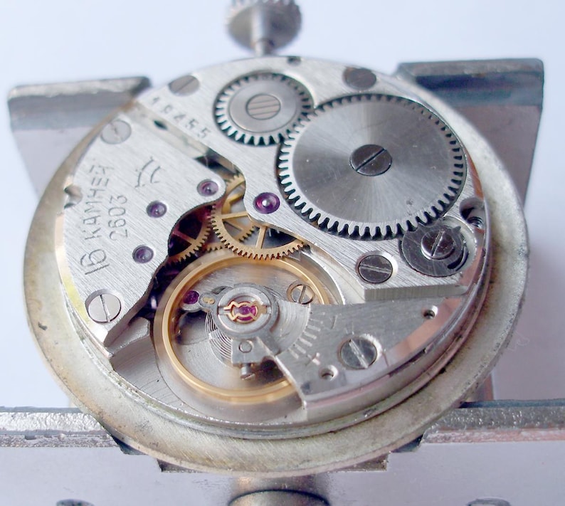 May include: Close-up of a disassembled watch movement. The silver-colored mechanism features numerous gears, screws, and jewels. The watch's internal components are visible, including the balance wheel and escapement. The watch movement is held in a silver metal frame.