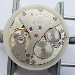 May include: A mechanical watch movement secured in a white plastic holder, featuring a silver-toned metal plate engraved with the text SEVENTEEN 17 JEWELS, visible gears, a balance wheel, and several small purple synthetic rubies throughout.