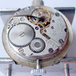 May include: Close-up of a watch movement with visible gears, screws, and other internal components. The watch mechanism is silver-toned with gold-colored gears and purple jewels. The text "16 камней 2605" is visible on the watch.