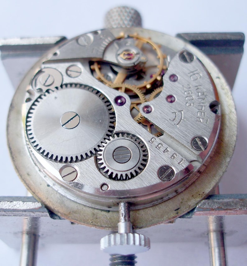 May include: Close-up of a watch movement with visible gears, screws, and other internal components. The watch mechanism is silver-toned with gold-colored gears and purple jewels. The text "16 камней 2605" is visible on the watch.