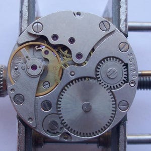 May include: A detailed view of a watch mechanism, showcasing intricate gears, screws, and a balance wheel. The metal parts are silver and gold, with the number "958200" visible. The watch movement is held in a metal tool.