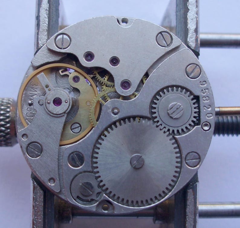 May include: A detailed view of a watch mechanism, showcasing intricate gears, screws, and a balance wheel. The metal parts are silver and gold, with the number "958200" visible. The watch movement is held in a metal tool.