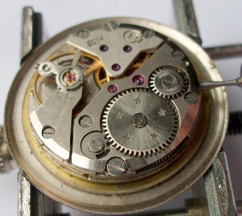 Pode incluir: Close-up de um movimento de rel&oacute;gio com engrenagens, parafusos e joias intrincadas. Os componentes met&aacute;licos s&atilde;o prateados e dourados, com um design circular. O rel&oacute;gio &eacute; preso em uma bra&ccedil;adeira de metal.