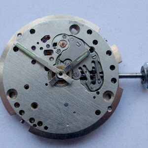May include: Close-up of a disassembled watch movement. The silver-colored mechanism features gears, screws, and a crown. The hands are black with green luminous paint. The watch movement is round and has several holes.