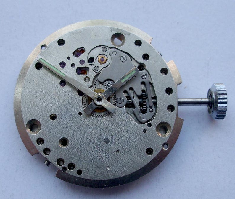 May include: Close-up of a disassembled watch movement. The silver-colored mechanism features gears, screws, and a crown. The hands are black with green luminous paint. The watch movement is round and has several holes.