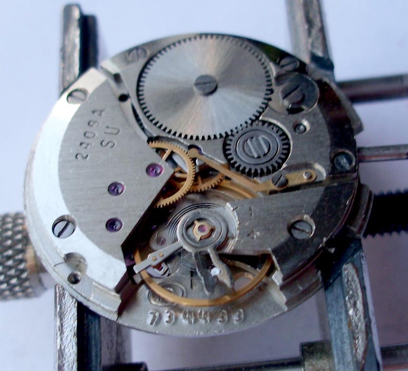 May include: Close-up of a watch movement with intricate gears and components. The silver-colored mechanism features multiple gears, screws, and jewels. The text "2409A SU" and "734433" are visible on the watch movement.