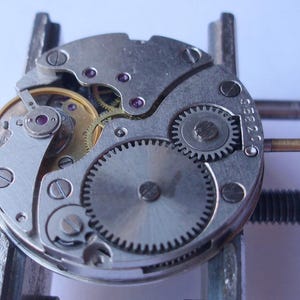 May include: Detailed view of a disassembled watch mechanism, revealing small gears, screws, and a gold-toned winding component. The silver-coloured parts are arranged in a circular design, with tiny ruby-coloured jewels adding a splash of colour. The watch is held in a metal clamp.