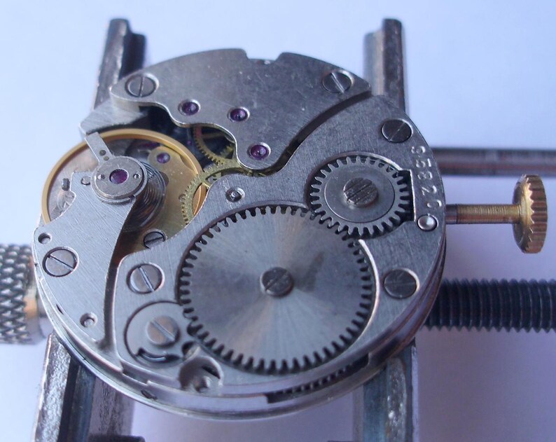 May include: Detailed view of a disassembled watch mechanism, revealing small gears, screws, and a gold-toned winding component. The silver-coloured parts are arranged in a circular design, with tiny ruby-coloured jewels adding a splash of colour. The watch is held in a metal clamp.