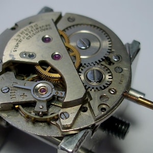 May include: Detailed view of a watch mechanism featuring silver and gold metal components, including gears, screws, and jewels. The watch movement has a gold crown stem and is labelled "PERRET SEVENTEEN JEWELS UNADJUSTED SWISS".
